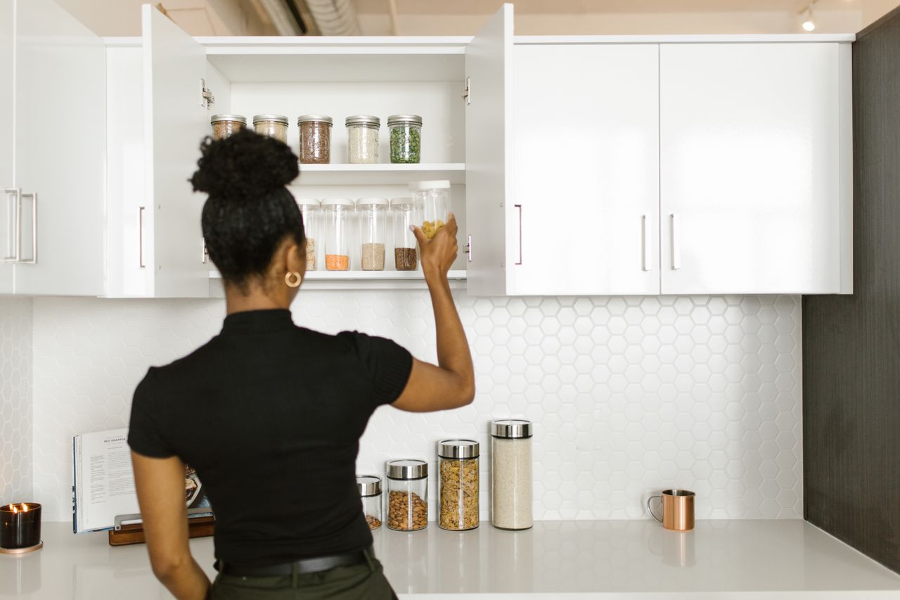 services-01 A woman organizing a pantry shelf with various food jars in a modern kitchen.